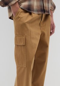 Helle Cargo-Hosen aus Baumwolle in Beige, mit einer Seitentasche und lockerer Passform, kombiniert mit einem karierten Hemd in neutralen Farbtönen.