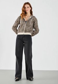 Hoodie s leopard uzorkom i bijelim detaljima, u kombinaciji s crnim širokim trapericama i crnim balerinkama na neutralnoj pozadini.