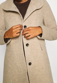 Abrigo de lana beige con un amplio cuello, cierre frontal de botones oscuros y bolsillos laterales. Tejido texturizado, diseño de longitud media.