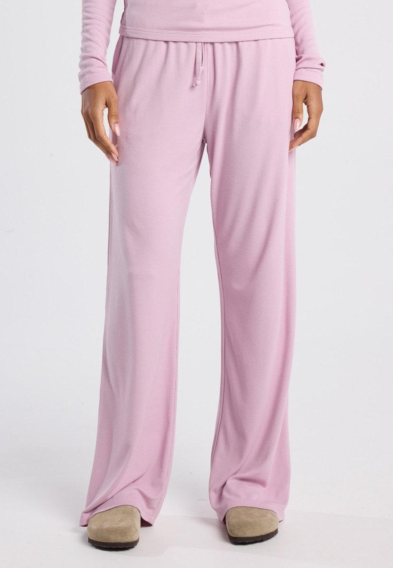 Light pink, loose-fitting, long pants made of soft fabric with an elastic waistband and drawstring. Pair of beige slippers visible.