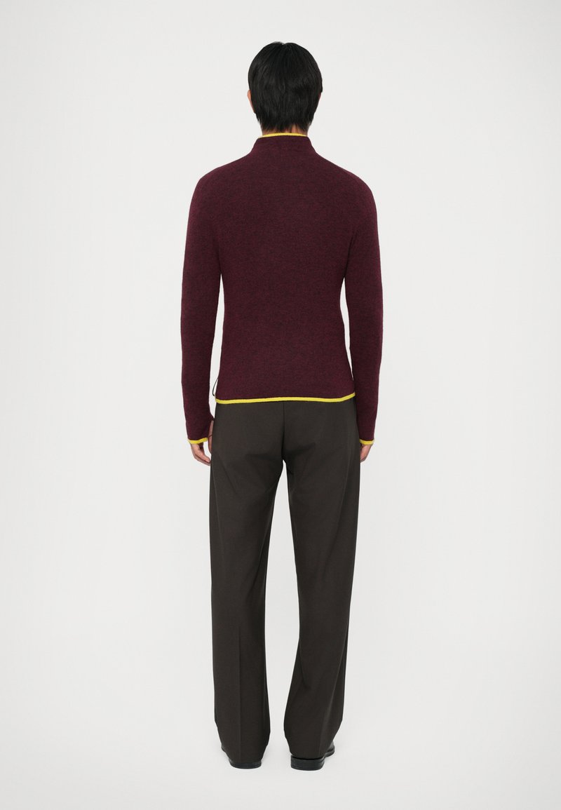 Bordeaux lange mouwen trui met gele afwerking bij de kraag en zoom, gecombineerd met donkere, losvallende broeken. Goedem tekstuur.