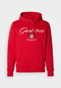 Sweatshirt à capuche rouge avec poche avant, cordons de serrage, et texte blanc "Gant 1949" au-dessus d'un emblème en forme de bouclier avec des étoiles et des rayures.