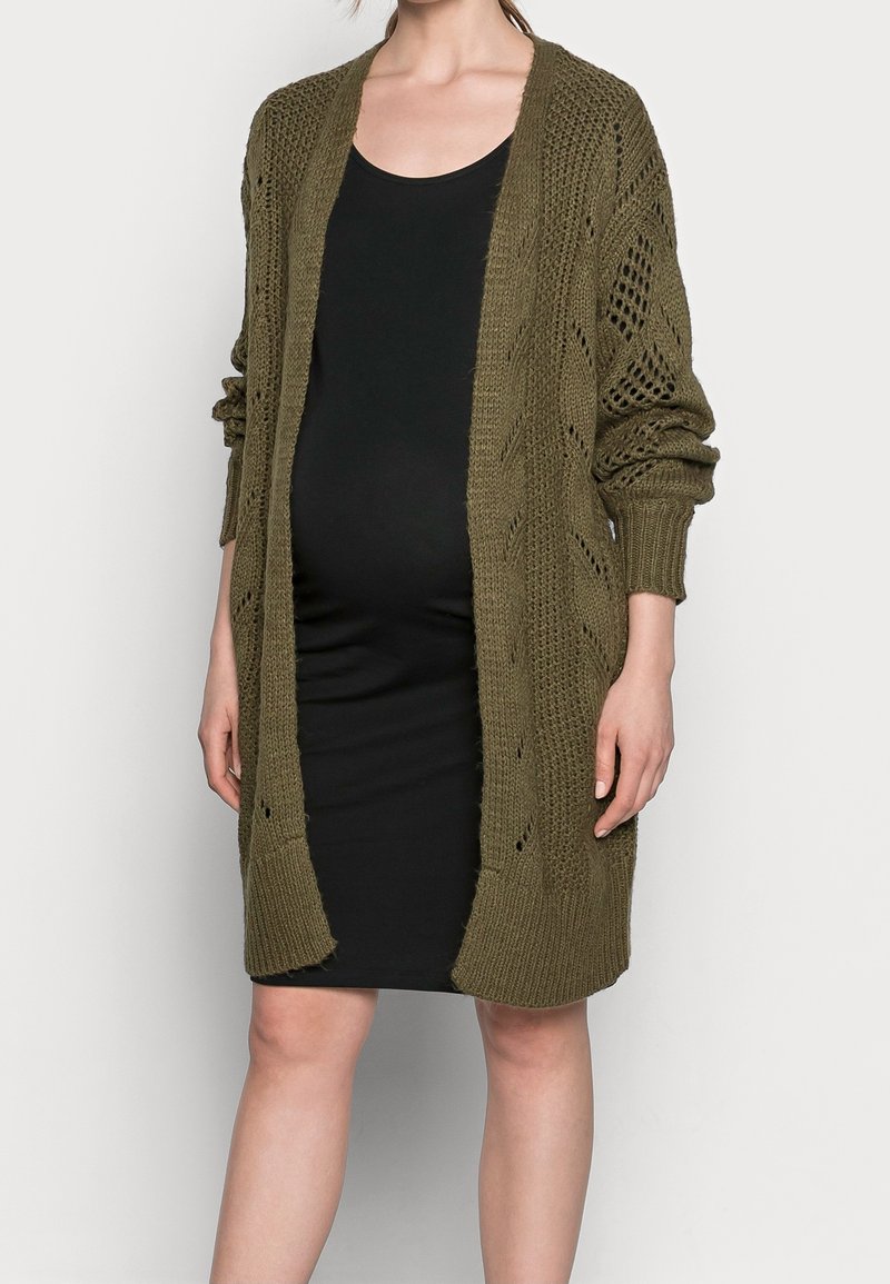 Olivově zelený pletený cardigan s otevřeným předním dílem, který má texturovaný vzor a ribbed manžety, vrstvený přes upnuté černé šaty.