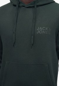 Άτομο που φοράει σκούρο πράσινο φούτερ Jack & Jones με κορδόνια, μπροστινή τσέπη και διακριτικό λογότυπο στο στήθος. Μόνο ο κορμός είναι ορατός.