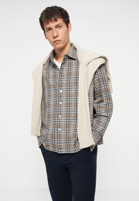 Brown plaid shirt with white and blue accents, button-down front, paired with a cream knit sweater draped over the shoulders.