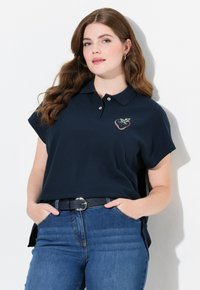 Polo shirt blu navy con maniche corte, caratterizzato da un ricamo di fragola sul petto. Abbinato a jeans blu e una cintura nera. Tessuto morbido.