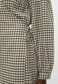 Musta ja beige ruudullinen mekko, jossa on solmitava yksityiskohta vyötäröllä ja tekstuuripintainen kangas. Pitkät, puffihihat, joissa on hupun yksityiskohta.
