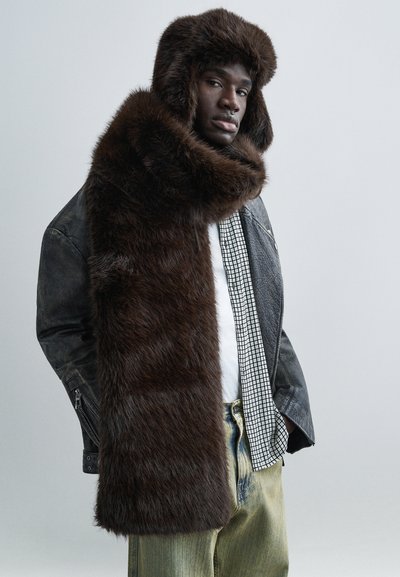 Άνδρας που φοράει υπερμεγέθη σκούφο από σκούρο καφέ γούνα και κασκόλ, μαύρο δερμάτινο μπουφάν, καρό πουκάμισο και ανοιχτόχρωμο μπλε τζιν, σε μονόχρωμο γκρίζο φόντο.