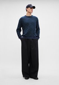 Marineblå strikket genser med strukturert diamantmønster, kombinert med vide svarte bukser og blanke svarte sko, samt en mørk caps.