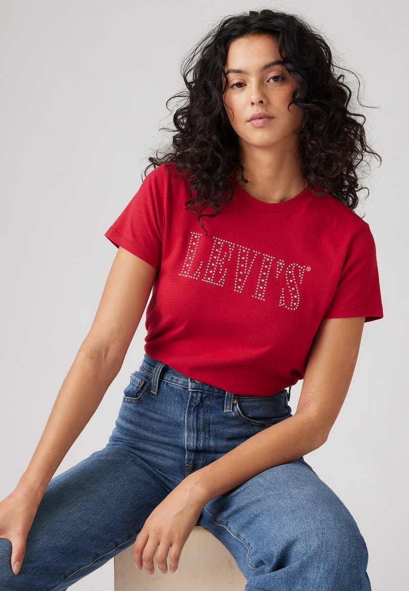Punane puuvillane T-särk lühikeste varrukatega, mille esiküljel on kividest kaunistatud "LEVI'S" logo. Kombineeritud kõrge vöökohaga siniste teksadega.