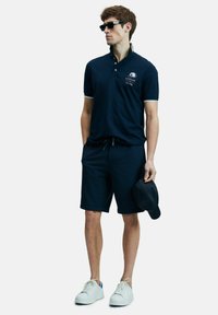 Man in een marineblauwe polo met bijpassende shorts, witte sneakers, een marineblauwe pet in de hand, met zwarte zonnebril, staande tegen een witte achtergrond.