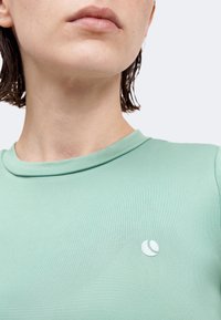 Κοντινό πλάνο ατόμου που φοράει πουκάμισο μέντας με μικρό λευκό ημισεληνοειδές λογότυπο στο στήθος, εμφανή τα χείλη και ο λαιμός.