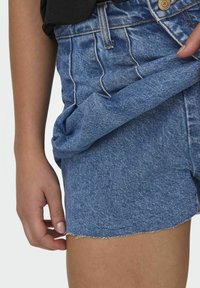 Denim kratke hlače v zbledeli modri barvi s strganim robom in vidno šivanko. Imajo visok pas in klasično zapiranje na gumb.