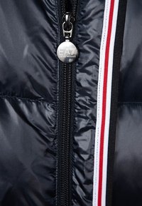 Černá lehká bunda s předním zipem, stříbrnými detaily a kontrastními bílými a červenými pruhy podél zipu.