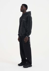 Black hoodie with front pocket and embroidered logo, paired with black sweatpants featuring a spider web pattern on the side, worn with black sneakers.