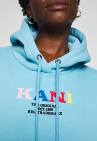 Sudadera azul claro con cordones. Presenta un colorido logo "KANI" en rosa, azul, rojo y amarillo. Material de algodón suave.