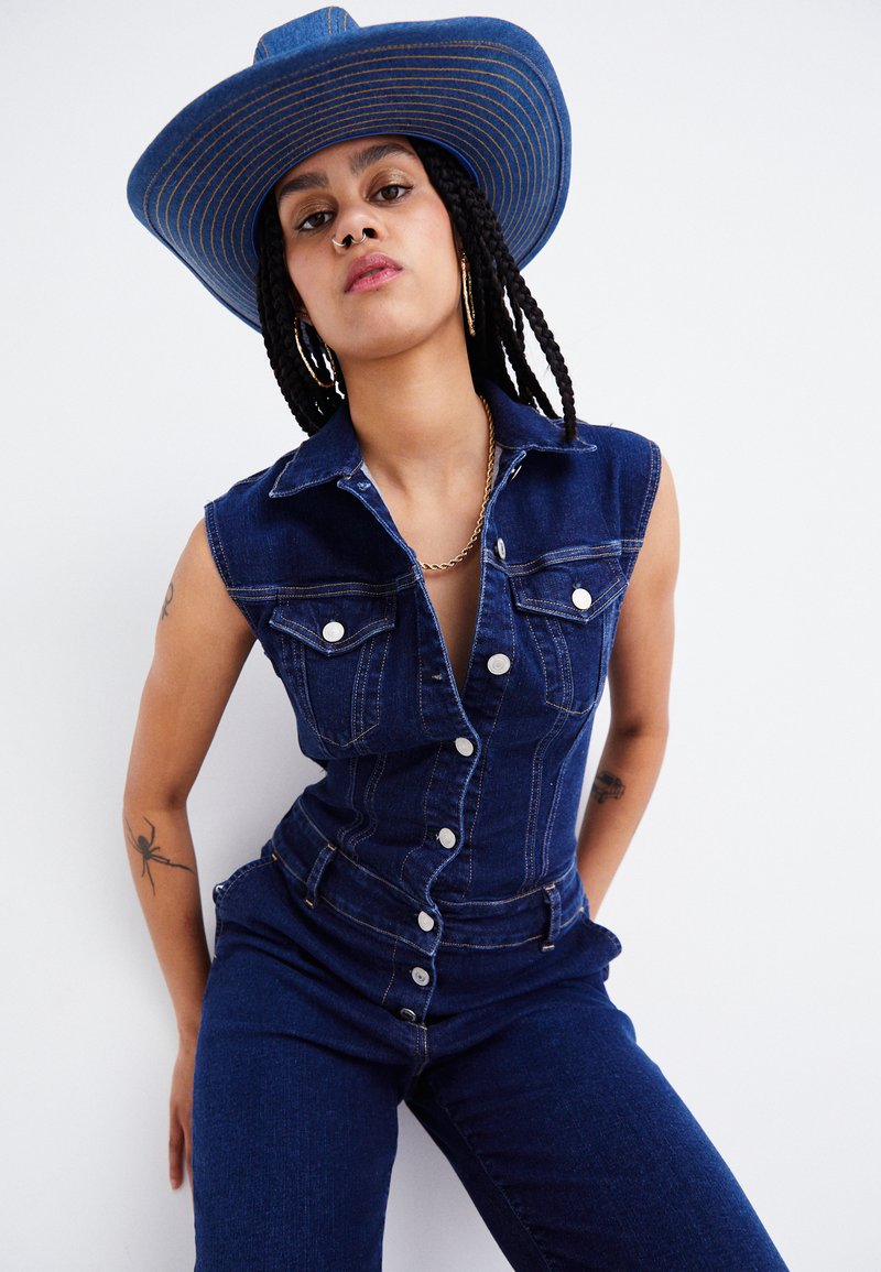 Salopette en denim avec un design sans manches, des poches frontales et une fermeture à boutons. Associée à un chapeau en denim à larges bords avec des accents rayés.