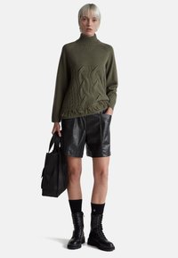 Olivově zelený pletený svetr s vysokým límcem a vzorem copánků, v kombinaci s černými koženými šortkami a černými combat botami. Součástí je černá taška.