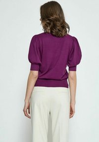 Lila gestrickter Pullover mit Puffärmeln und geripptem Saum, kombiniert mit hellbeigen, weiten Hosen, gezeigt von hinten.