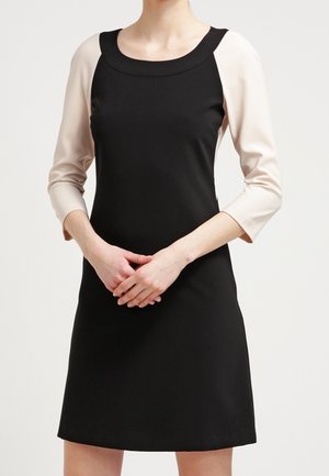 Frau trägt ein schwarzes ärmelloses Kleid über einem beigen Langarmshirt und steht mit verschränkten Händen vor sich.