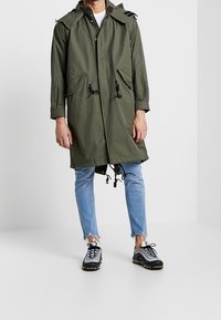 Impermeabile lunghe in verde oliva con cappuccio, dotato di due tasche frontali e cordini in vita e sull'orlo. Indossato con jeans azzurri e sneakers.