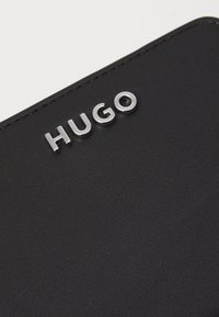Portefeuille en cuir noir avec une surface texturée et le logo "HUGO" embossé en métal argenté. Caractéristiques : lignes épurées et design minimaliste.