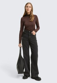 Braun gepunkteter Rollkragenpullover, hochsitzzende schwarze weite Jeans und schwarze klobige Schuhe. Hält eine schwarze Tasche.