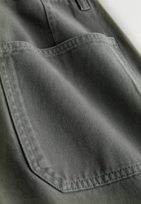 Zbliżenie szarej kieszeni z tkaniny z wzmocnionym podwójnym przeszyciem na elemencie odzieży, ukazujące szczegółową fakturę i szwy.