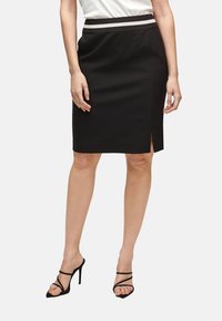 Černá tužková sukně se bočním rozparkem, s kontrastním pruhovaným pasem. Hladká tkanina s klasickým střihem, nošena s černými sandály na vysokém podpatku.