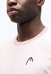 Κοντινό πλάνο του κάτω μέρους του προσώπου και του λαιμού ενός ατόμου που φοράει ένα ανοιχτό ροζ πουκάμισο με μικρό σκούρο λογότυπο στο στήθος.