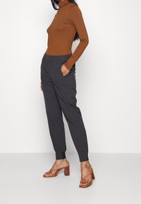 Hnědý dlouhý rukáv turtleneck svetr zkombinovaný se šedými upravenými kalhotami s bočními kapsami, tapered nohavicemi a černými kotníkovými členkovými manžetami. Hnědé sandály na podpatku.