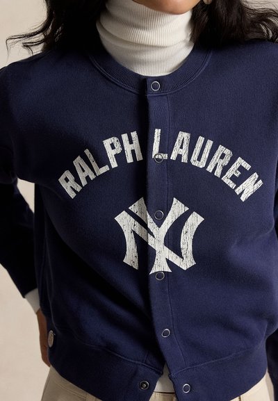 Άτομο φοράει ένα σκούρο μπλε φούτερ Ralph Lauren με κουμπιά πάνω από ένα λευκό ζιβάγκο, εμφανίζοντας το όνομα και το λογότυπο της μάρκας στο στήθος.