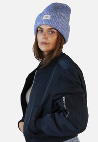Light blue knitted beanie with ribbed texture and sewn brand label. Paired with a black bomber jacket featuring zippered pockets.