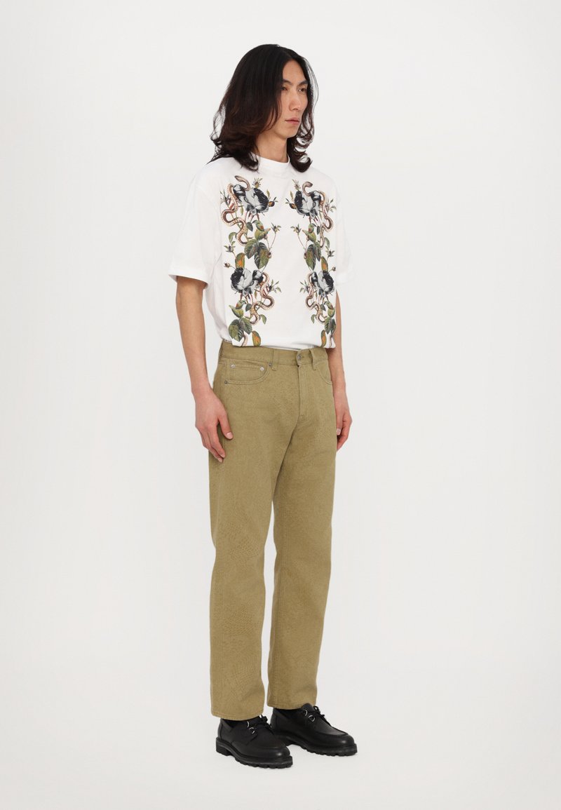 T-shirt bianco con stampa botanica e animale, abbinato a pantaloni di velluto a coste verde oliva chiaro. Scarpe nere con lacci completano il look.