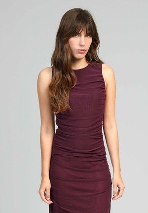 Femme aux longs cheveux bruns portant une robe sans manches froncée de couleur violet foncé, se tenant devant un fond gris clair uni.