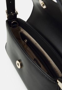 Bolso de mano de cuero negro con textura lisa, herrajes en tono dorado y un bolsillo con cremallera en el interior sobre un forro beige con patrón de logotipos.