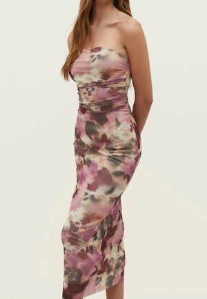 Femme portant une robe midi moulante sans bretelles avec un motif floral abstrait rose, marron et beige, debout devant un fond uni.