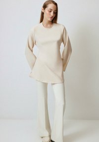 Beige long-sleeve top with a round neckline, smooth texture, and relaxed fit, paired with white flared pants and slip-on shoes.