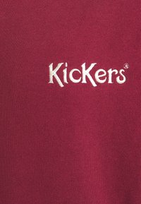 Weiß gesticktes "Kickers"-Logo auf einer Nahaufnahme von strukturiertem burgunderrotem Stoff, wahrscheinlich ein Bekleidungsstück.