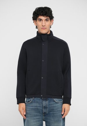 Svart jakke laget av myk stoff, med stående krage, trykknapper og sidelommer. Kombinert med lyseblå denimjeans.