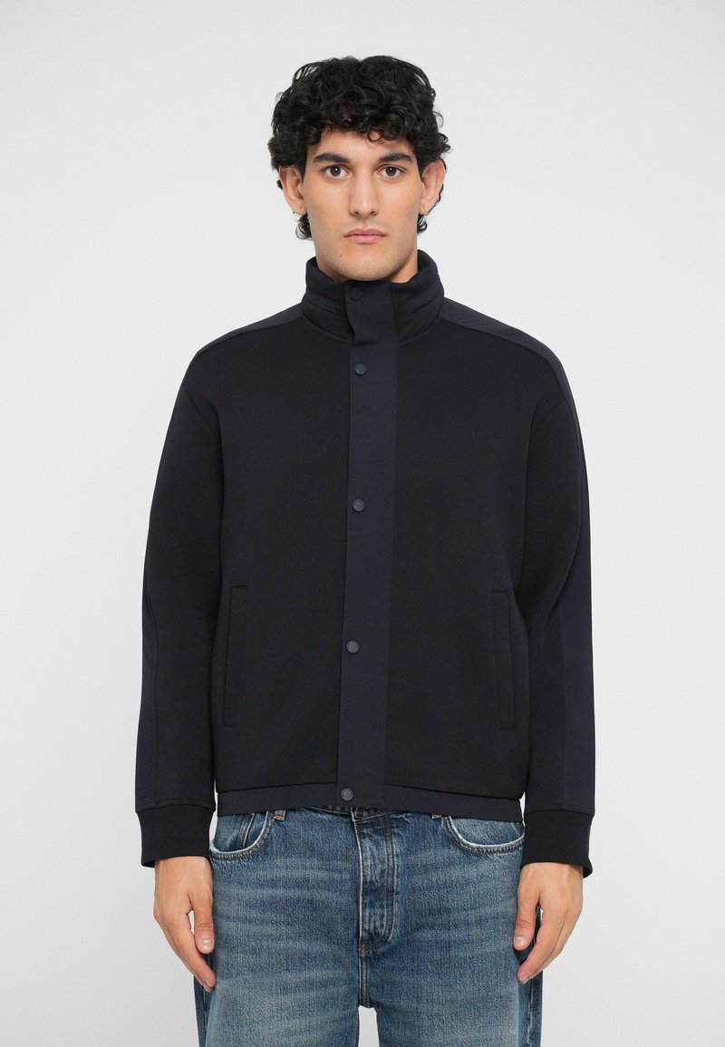 Veste noire en tissu doux, dotée d'un col montant, de boutons-pression et de poches latérales. Associée à un jean en denim bleu clair.