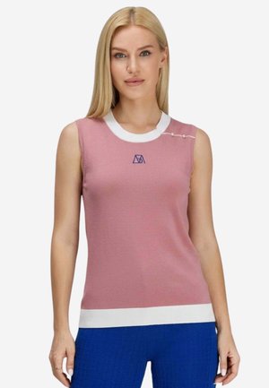 Donna bionda con top rosa senza maniche con bordo bianco e pantaloni blu, in piedi davanti a uno sfondo chiaro e uniforme.