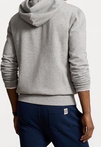 Felpa grigia con cappuccio, maniche lunghe, polsini e orlo a coste. Indossata con pantaloni blu scuro con un'etichetta sulla tasca posteriore.