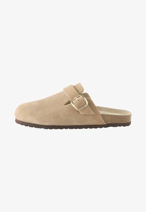 Scarpa slip-on in suede di colore beige con punta arrotondata, una cinghia con fibbia sulla parte superiore e una suola marrone testurizzata.