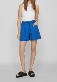Blue high-waisted shorts with a tied belt, side pockets, and a relaxed fit. Paired with a fitted white sleeveless top and black sandals.