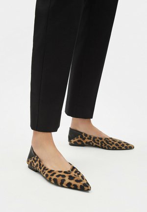Nogi w czarnych spodniach i czarne baleriny w cętki lamparta z czarnymi nasadkami na obcasach na białym tle.