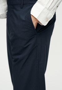 Χέρι στην τσέπη σκούρου ναυτικού μπλε παντελονιού που φοριέται με λευκό πουκάμισο μακρυμάνικο με κουμπωτό μανίκι, ορατό από τη μέση μέχρι τον μηρό.