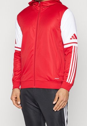 Personne portant une veste de sport rouge et blanche zippée avec capuche et un pantalon noir avec des bandes blanches sur le côté.