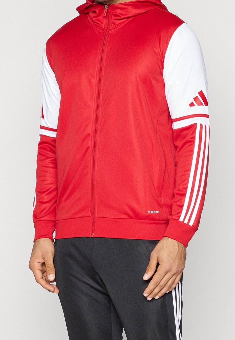 Personne portant une veste de sport rouge et blanche zippée avec capuche et un pantalon noir avec des bandes blanches sur le côté.