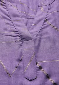 Lila Stoffoberteil mit V-Ausschnitt-Design, das goldene abstrakte Muster und eine Taschendetail aufweist. Glatte Textur mit subtiler Glanz.